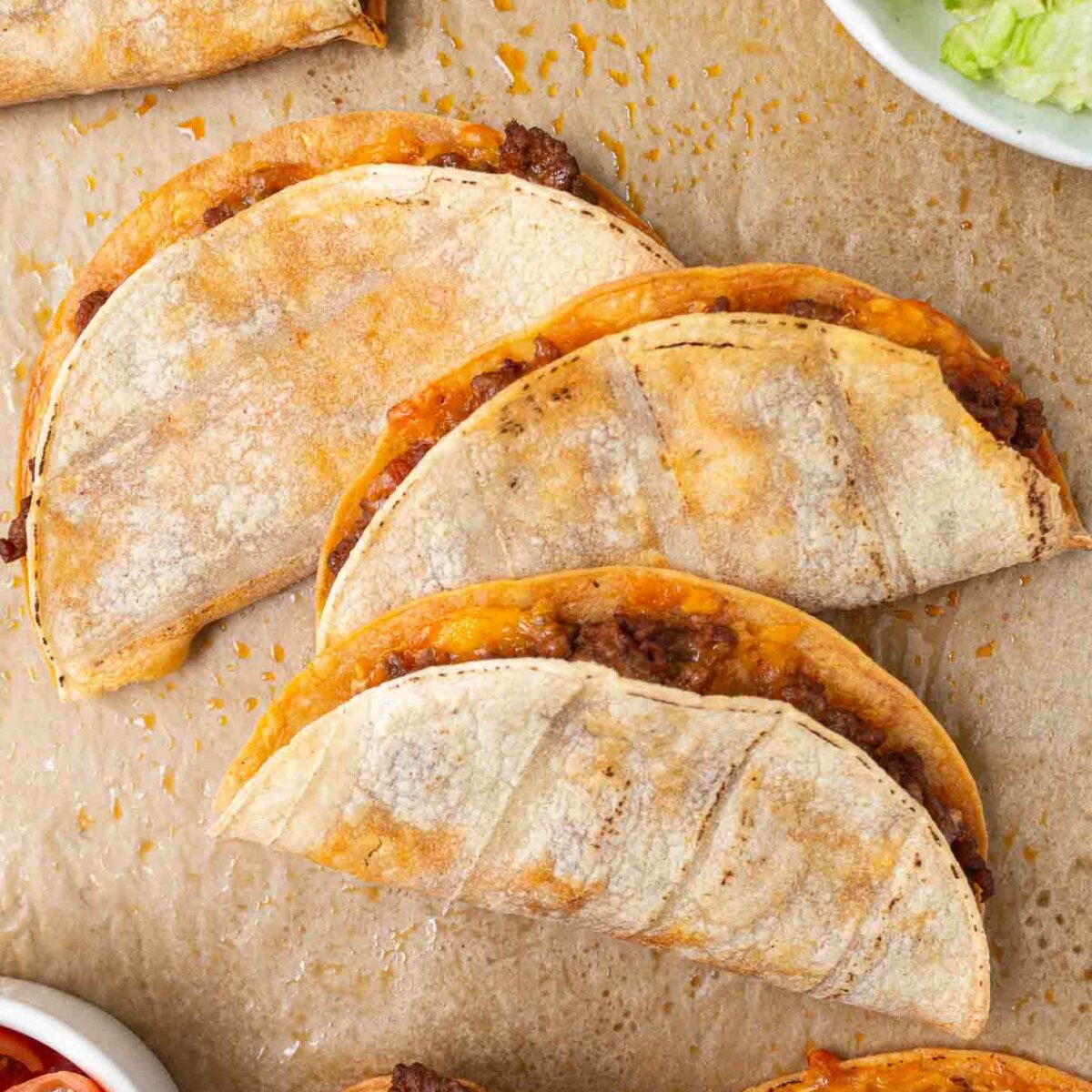 Close-up of crispy tacos filled with seasoned ground meat and cheese, placed on a parchment-lined surface. Bowls of chopped tomatoes and shredded lettuce are on the side, suggesting toppings for the tacos.