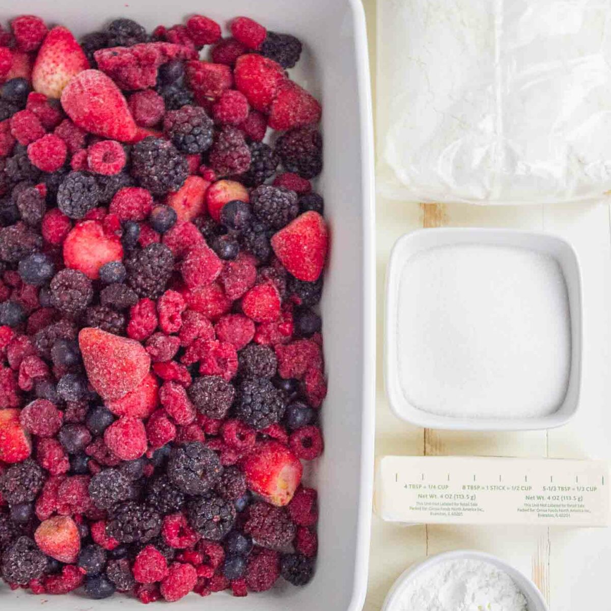 A white dish filled with mixed frozen berries sits next to a stick of butter, a small bowl of sugar, a bowl of cornstarch, and a plastic bag of cake mix on a light wooden surface.