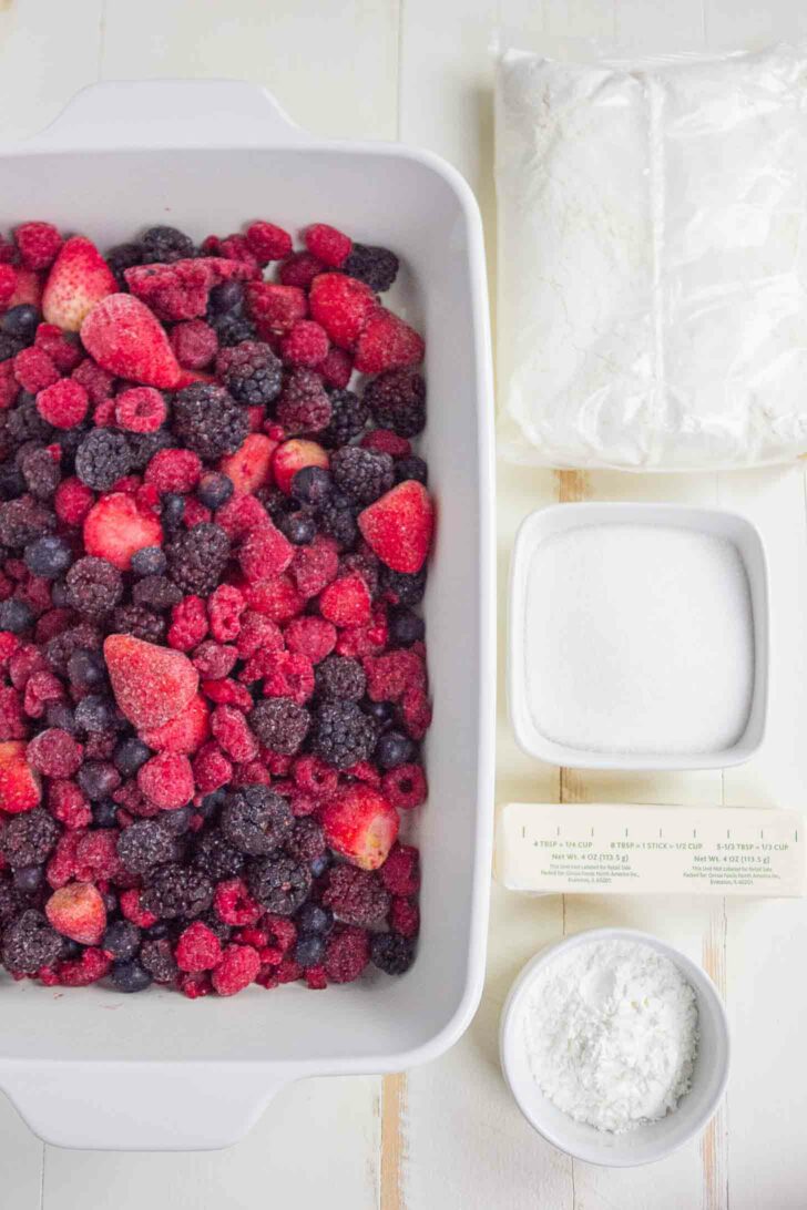 A white dish filled with mixed frozen berries sits next to a stick of butter, a small bowl of sugar, a bowl of cornstarch, and a plastic bag of cake mix on a light wooden surface.