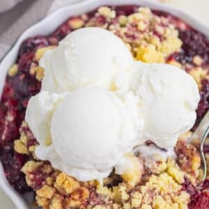 A close-up of a berry cobbler dessert topped with three scoops of vanilla ice cream in a white dish. The crumble appears golden and crisp, with visible baked berries underneath.