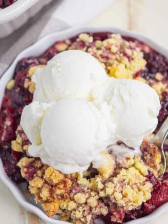 A close-up of a berry cobbler dessert topped with three scoops of vanilla ice cream in a white dish. The crumble appears golden and crisp, with visible baked berries underneath.