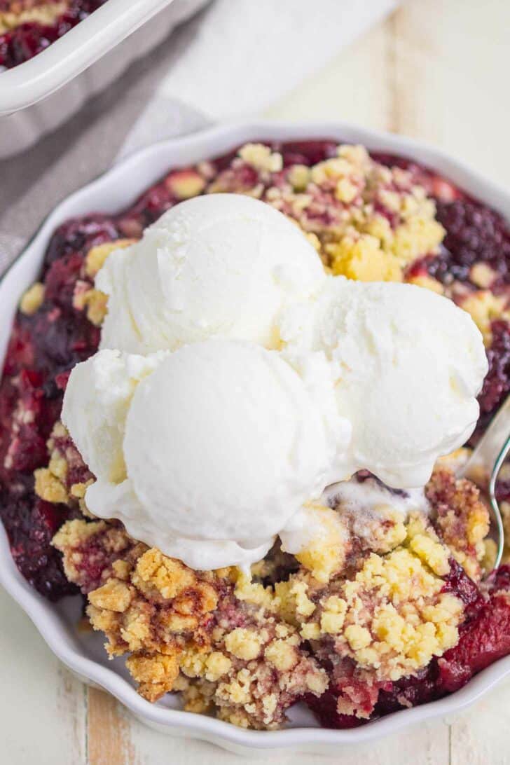 A close-up of a berry cobbler dessert topped with three scoops of vanilla ice cream in a white dish. The crumble appears golden and crisp, with visible baked berries underneath.