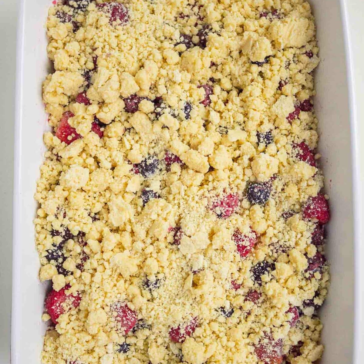 A white baking dish filled with an unbaked berry cobbler, showing a layer of mixed berries topped with a crumbly, yellow cake mix