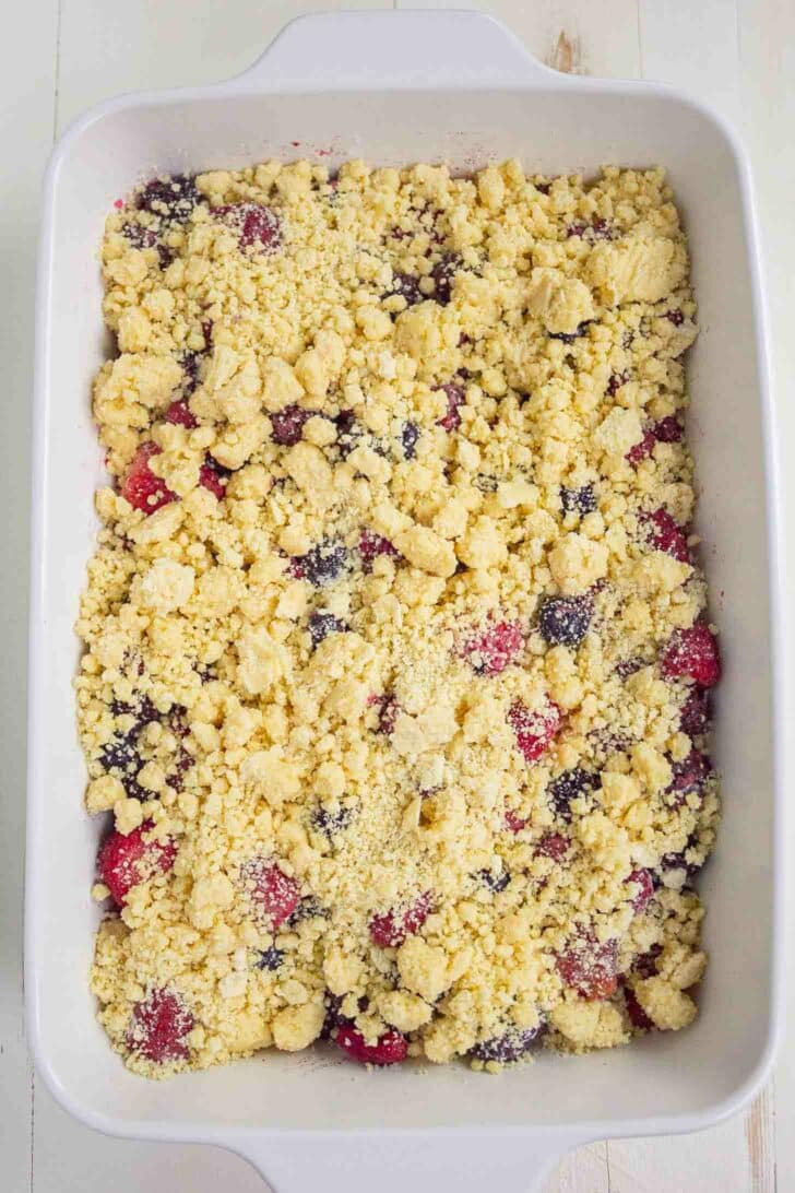 A white baking dish filled with an unbaked berry cobbler, showing a layer of mixed berries topped with a crumbly, yellow cake mix