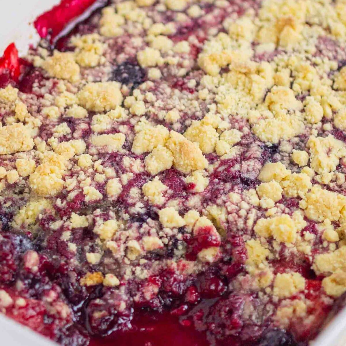 A close-up of a baked berry cobbler in a white dish, showing bubbling mixed berries and a golden, crumbly topping.