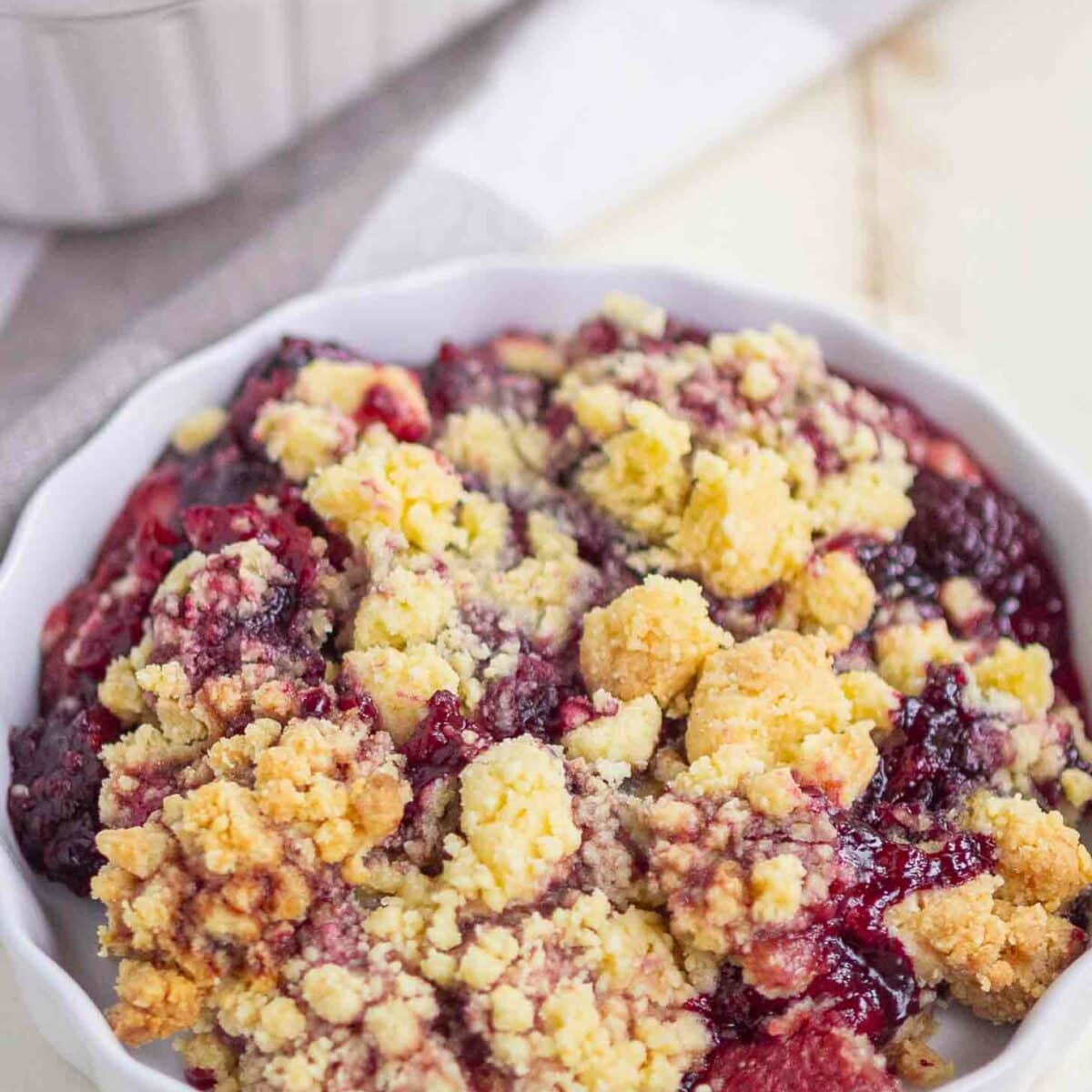 A close-up of a small white dish filled with berry cobbler, featuring a golden, crumbly topping and vibrant mixed berry filling. A larger baking dish with more crumble is visible in the background.