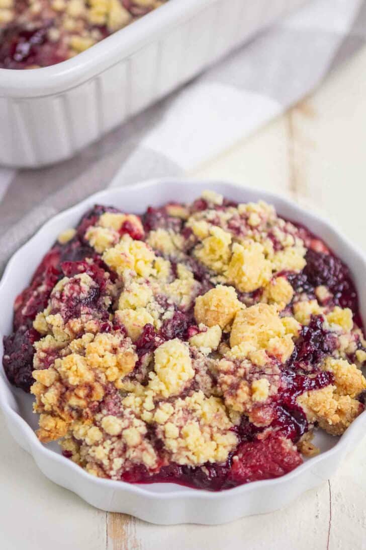 A close-up of a small white dish filled with berry cobbler, featuring a golden, crumbly topping and vibrant mixed berry filling. A larger baking dish with more crumble is visible in the background.