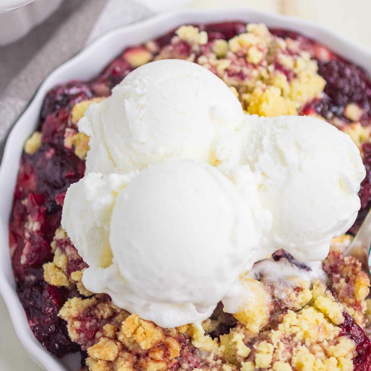 A serving of berry cobbler topped with three scoops of vanilla ice cream in a white dish, showing a mix of golden crumble and juicy berries underneath.