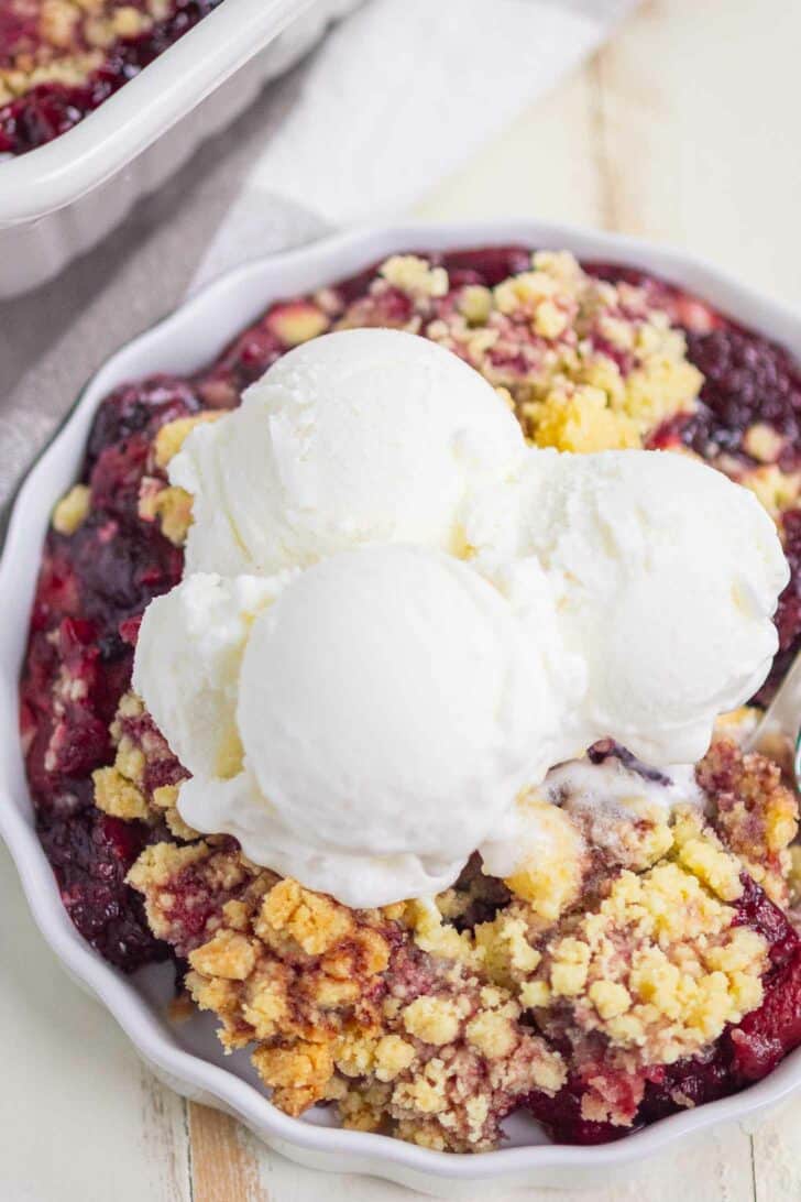A serving of berry cobbler topped with three scoops of vanilla ice cream in a white dish, showing a mix of golden crumble and juicy berries underneath.