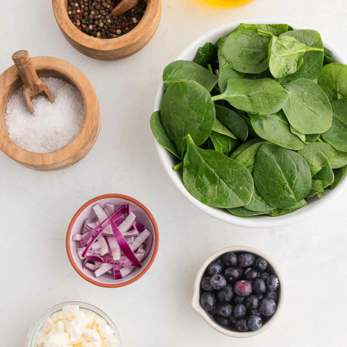 A bowl of fresh spinach leaves is surrounded by small bowls of salt, pepper, olive oil, sliced red onions, blueberries, crumbled cheese, and balsamic vinegar on a light countertop.
