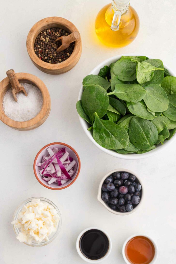 A bowl of fresh spinach leaves is surrounded by small bowls of salt, pepper, olive oil, sliced red onions, blueberries, crumbled cheese, and balsamic vinegar on a light countertop.