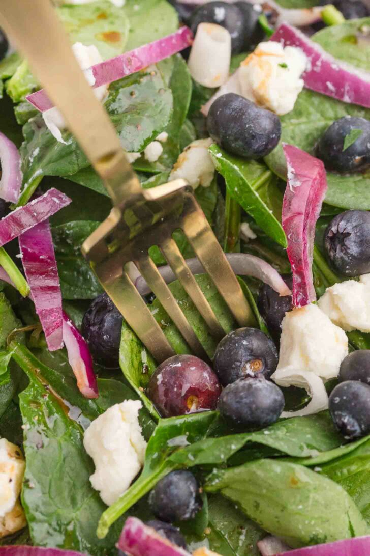 A close-up of a fresh salad featuring spinach leaves, blueberries, red onion slices, crumbled feta cheese, and a single grape. A gold fork is visible in the center, piercing the ingredients.