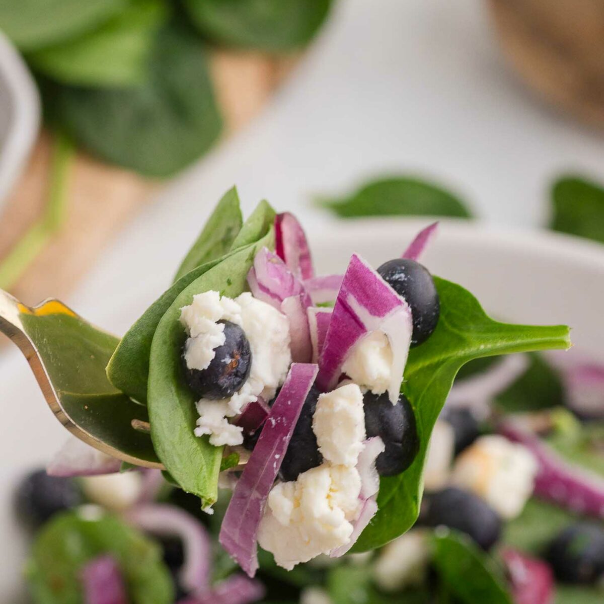 A close-up of a fresh salad on a fork, featuring spinach leaves, blueberries, red onion slices, and crumbled feta cheese. The rest of the salad is blurred in the background, and a bowl of spinach is partially visible.