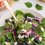 A fresh salad featuring spinach leaves, blueberries, crumbled feta cheese, and sliced red onions is being mixed with a gold fork in a white bowl. A wooden bowl with salt and fresh spinach leaves are visible in the background.