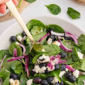 A fresh salad featuring spinach leaves, blueberries, crumbled feta cheese, and sliced red onions is being mixed with a gold fork in a white bowl. A wooden bowl with salt and fresh spinach leaves are visible in the background.