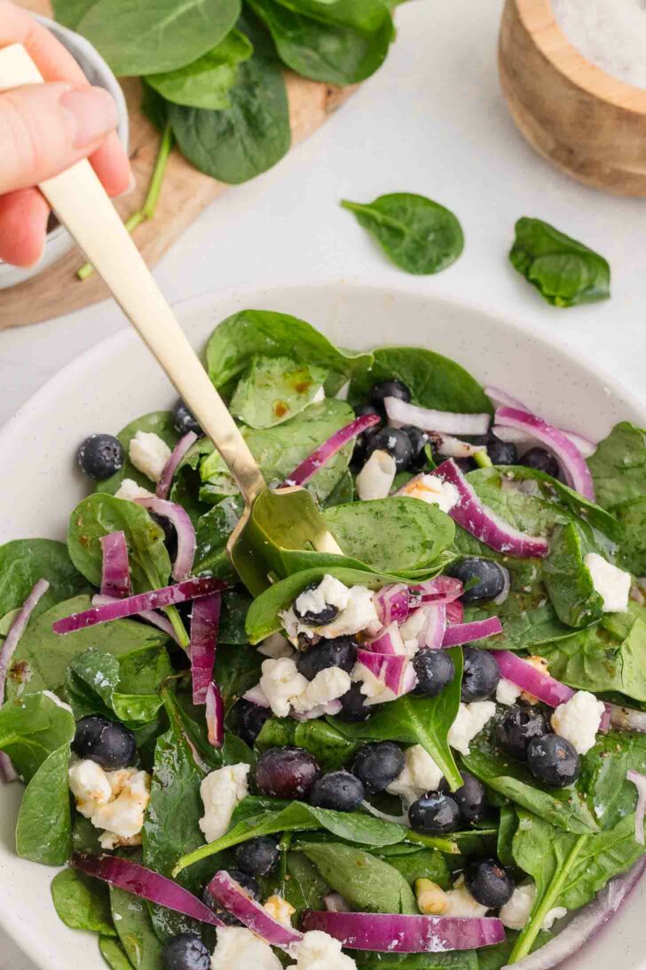 A fresh salad featuring spinach leaves, blueberries, crumbled feta cheese, and sliced red onions is being mixed with a gold fork in a white bowl. A wooden bowl with salt and fresh spinach leaves are visible in the background.