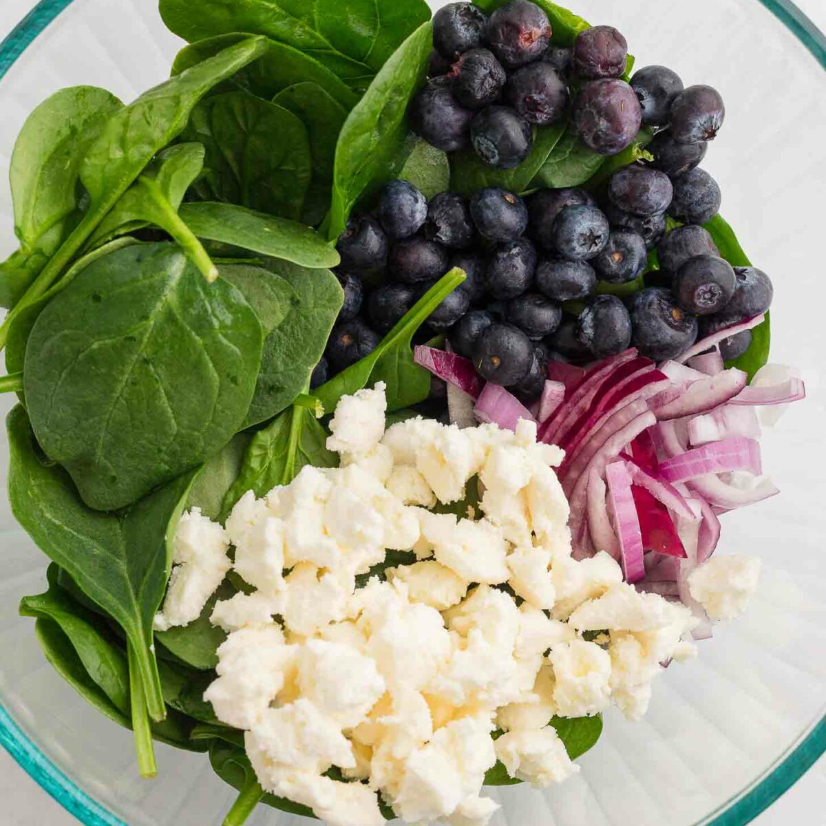 A glass bowl containing fresh spinach leaves, blueberries, sliced red onions, and crumbled cheese, arranged separately. The salad ingredients are vibrant and colorful against a light background.