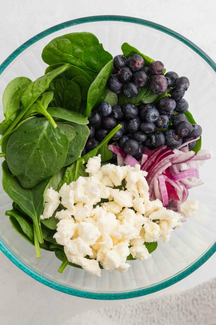 A glass bowl containing fresh spinach leaves, blueberries, sliced red onions, and crumbled cheese, arranged separately. The salad ingredients are vibrant and colorful against a light background.