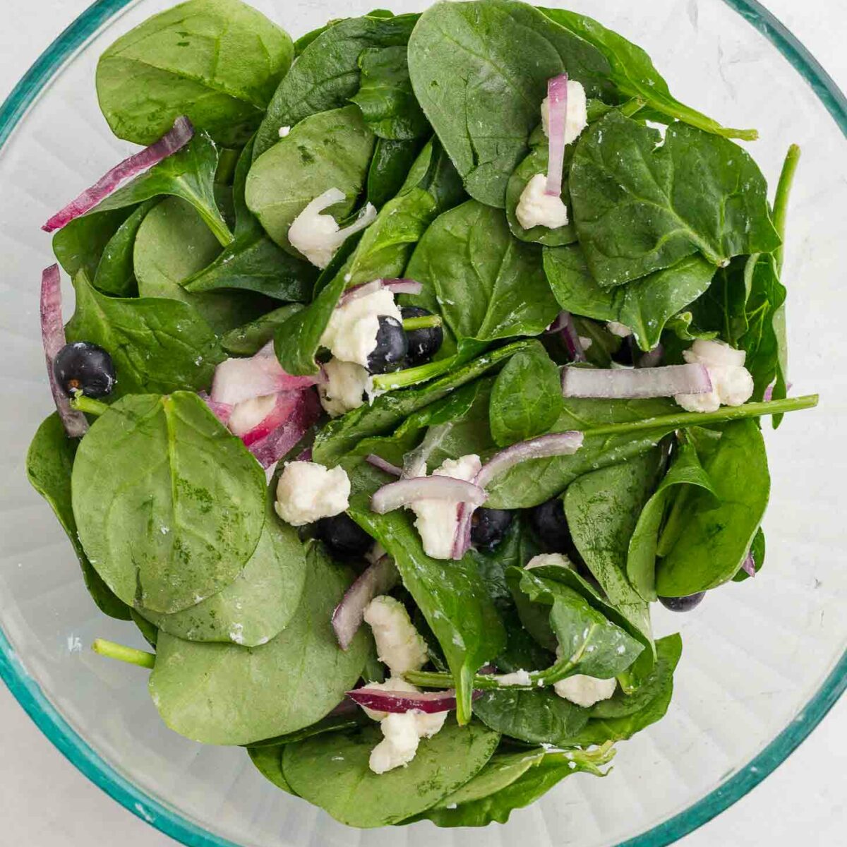 A glass bowl filled with fresh spinach leaves, slices of red onion, blueberries, and crumbles of white cheese, all lightly dressed, set against a light background.