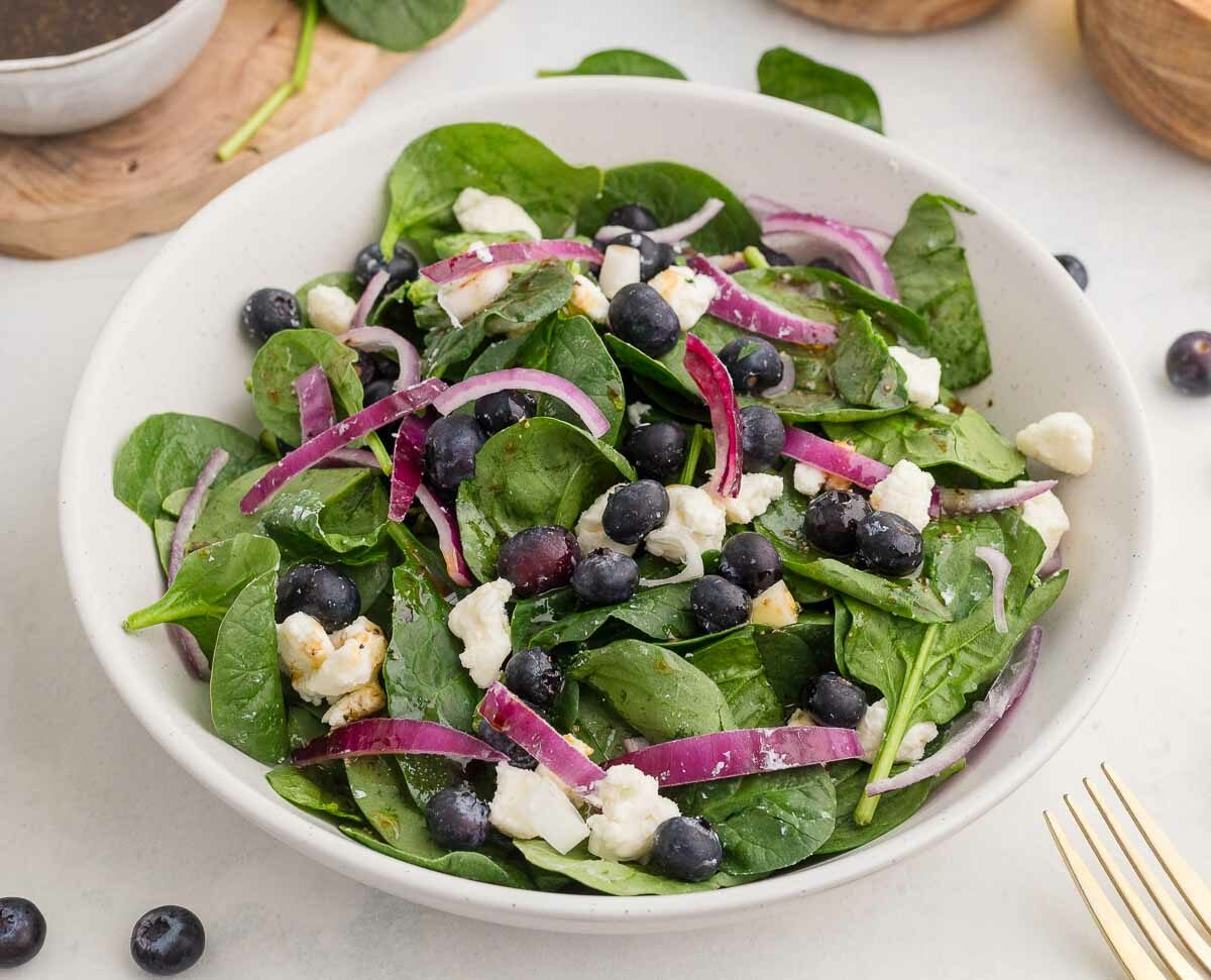 A bowl of fresh salad containing spinach leaves, blueberries, red onion slices, and crumbled feta cheese. A dressing container and wooden bowls are in the background. Forks and a striped napkin are placed beside the bowl.