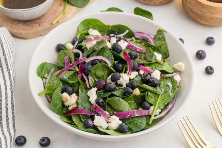 A bowl of fresh salad containing spinach leaves, blueberries, red onion slices, and crumbled feta cheese. A dressing container and wooden bowls are in the background. Forks and a striped napkin are placed beside the bowl.