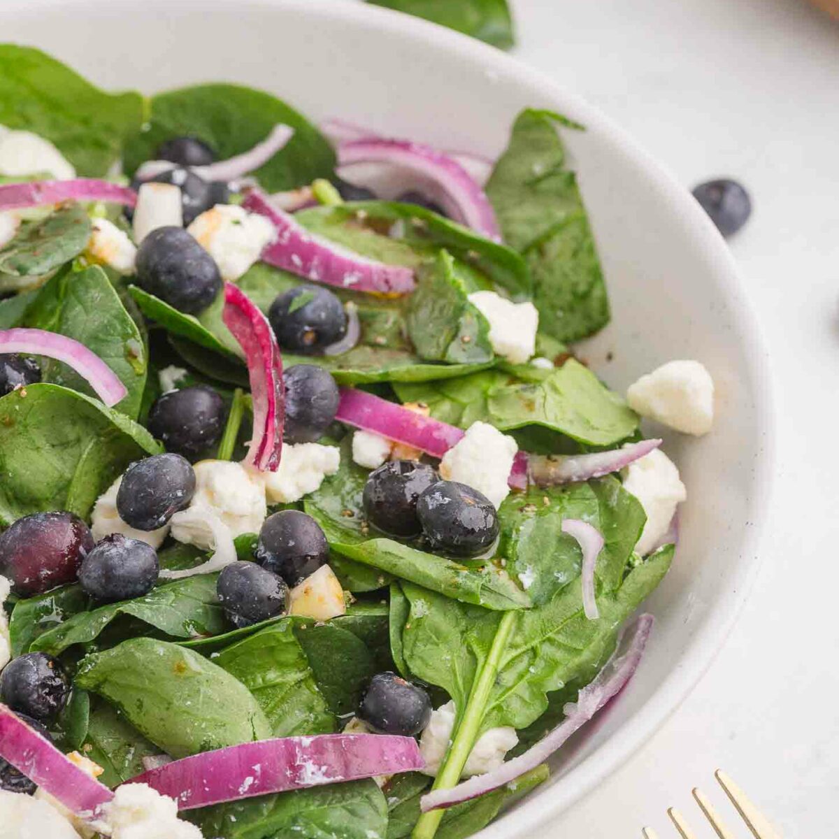 A fresh salad in a white bowl featuring spinach leaves, blueberries, crumbled feta cheese, and sliced red onions. A gold fork is partially visible on the side, and the background shows wooden bowls.