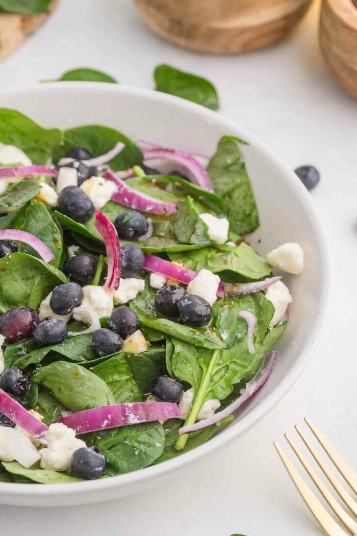 A fresh salad in a white bowl featuring spinach leaves, blueberries, crumbled feta cheese, and sliced red onions. A gold fork is partially visible on the side, and the background shows wooden bowls.