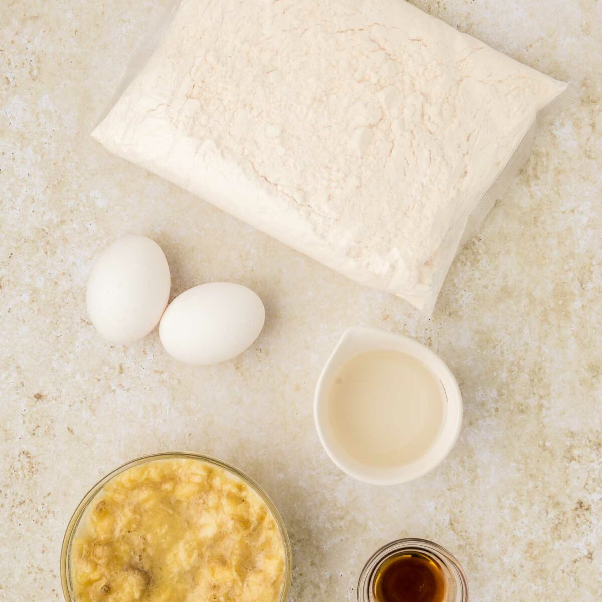 Ingredients for cake mix banana bread: a bag of cake mix, two eggs, a small bowl of mashed bananas, a cup of milk, and a small dish of vanilla extract on a light textured surface.