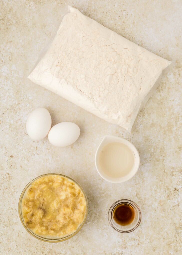 Ingredients for cake mix banana bread: a bag of cake mix, two eggs, a small bowl of mashed bananas, a cup of milk, and a small dish of vanilla extract on a light textured surface.