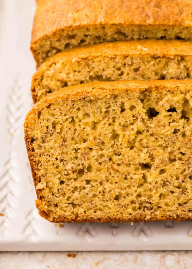 Close-up of sliced cake mix banana bread on a white rectangular plate. The bread has a golden-brown crust and a soft, moist interior with visible banana speckles, showcasing its fluffy texture.