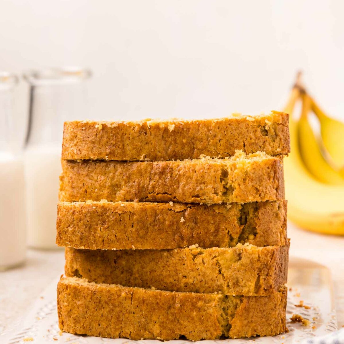 A stack of five slices of cake mix banana bread on a white cutting board. In the background, there are two glass bottles of milk and some bananas. A striped cloth is partially visible in the foreground.