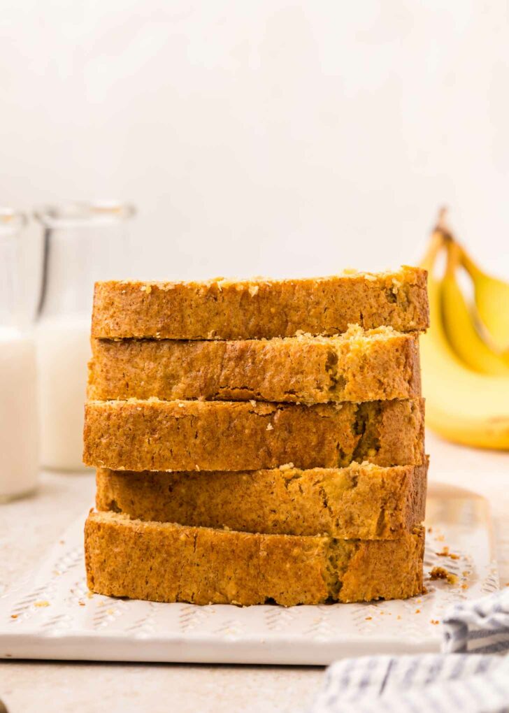 A stack of five slices of cake mix banana bread on a white cutting board. In the background, there are two glass bottles of milk and some bananas. A striped cloth is partially visible in the foreground.