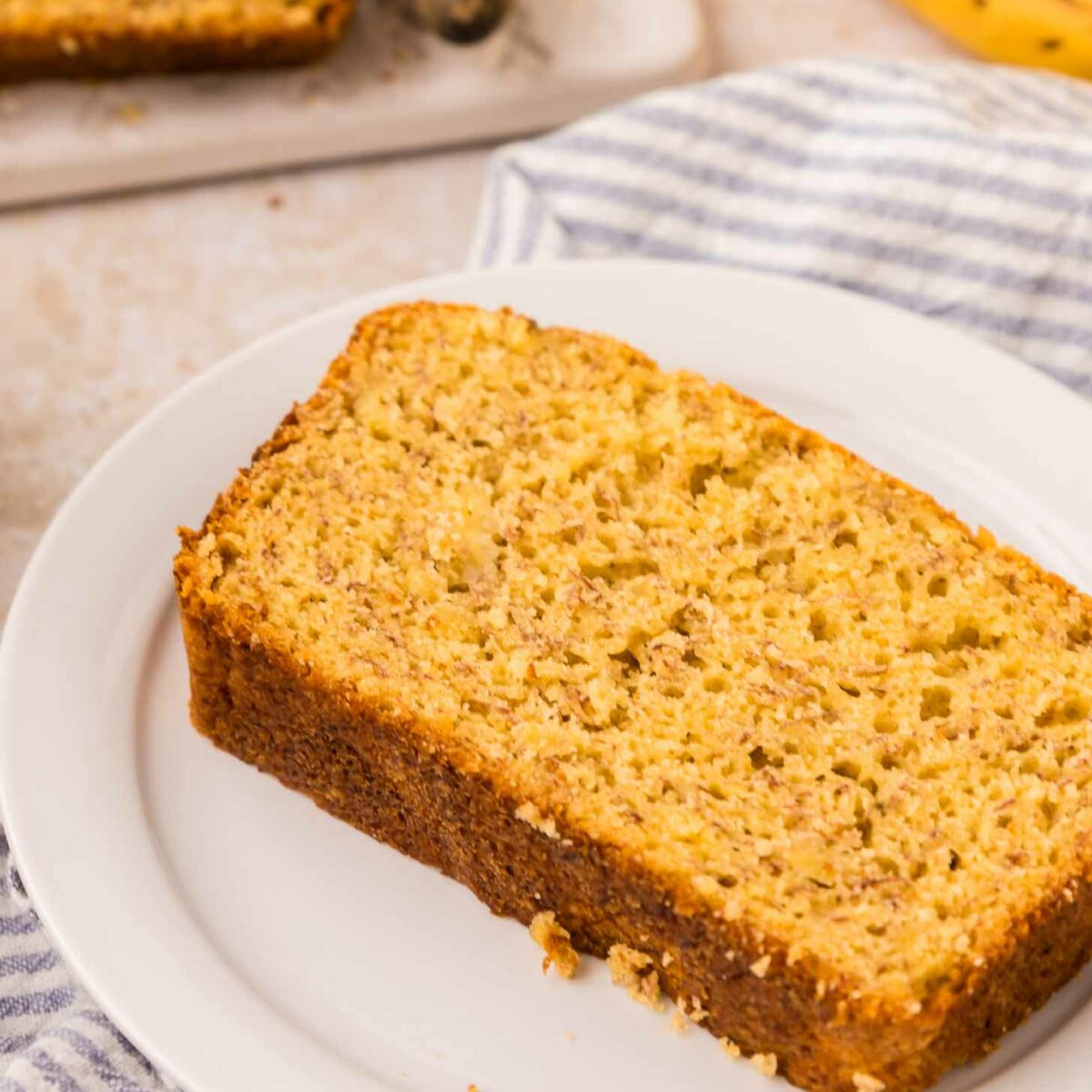 A slice of cake mix banana bread on a white plate, set on a striped cloth. A whole banana and more sliced bread are visible in the background, all on a light-colored surface.