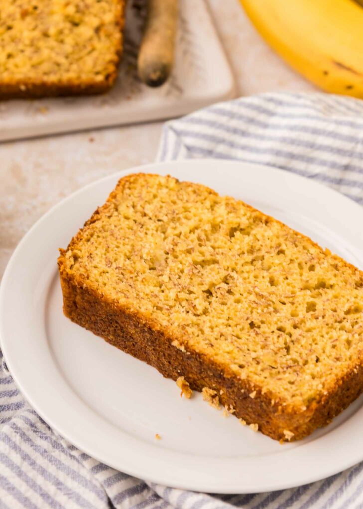 A slice of cake mix banana bread on a white plate, set on a striped cloth. A whole banana and more sliced bread are visible in the background, all on a light-colored surface.