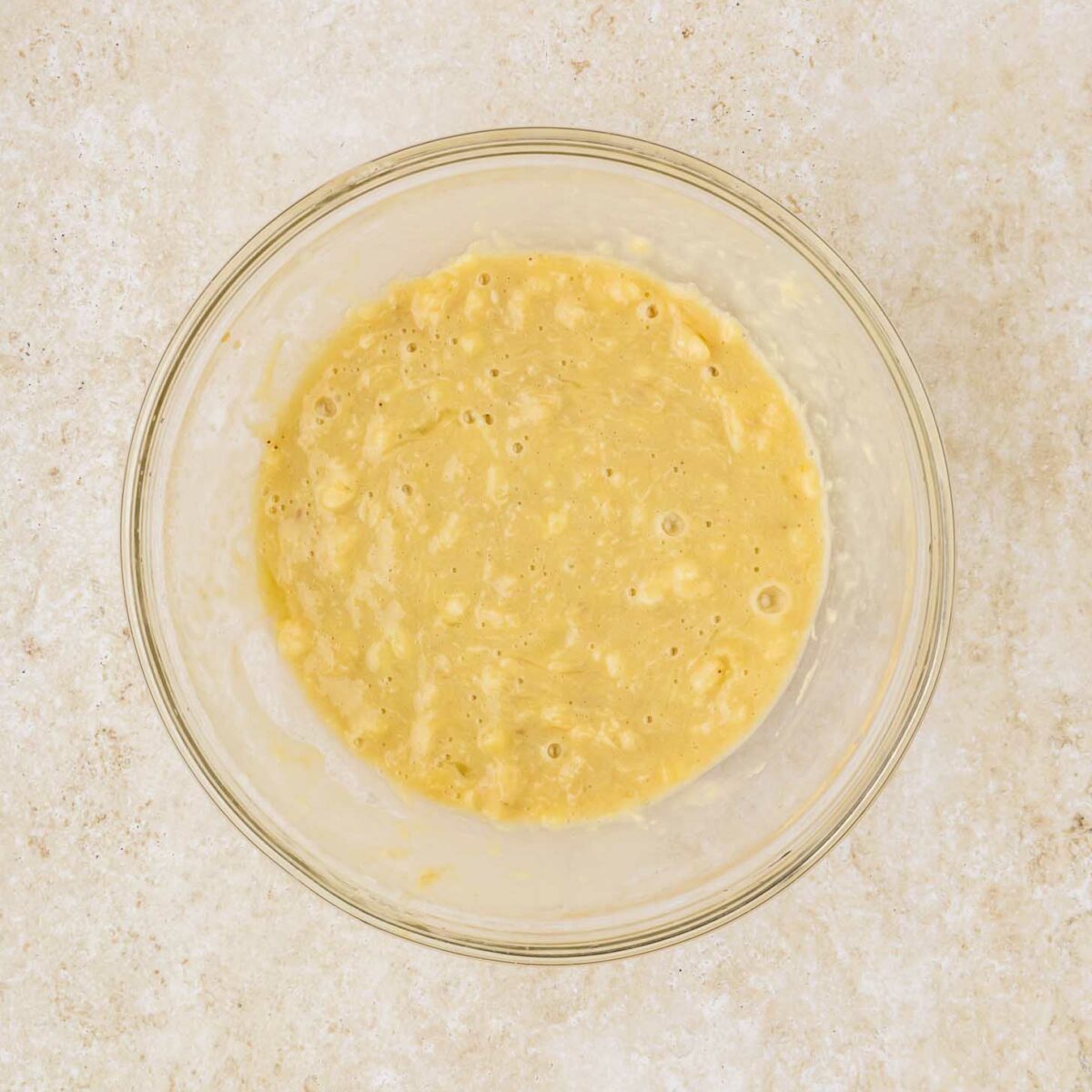 A clear glass bowl filled with a smooth, creamy cake mix banana bread batter rests on a light, speckled countertop. The batter is pale yellow and slightly lumpy, indicating the presence of mashed bananas.