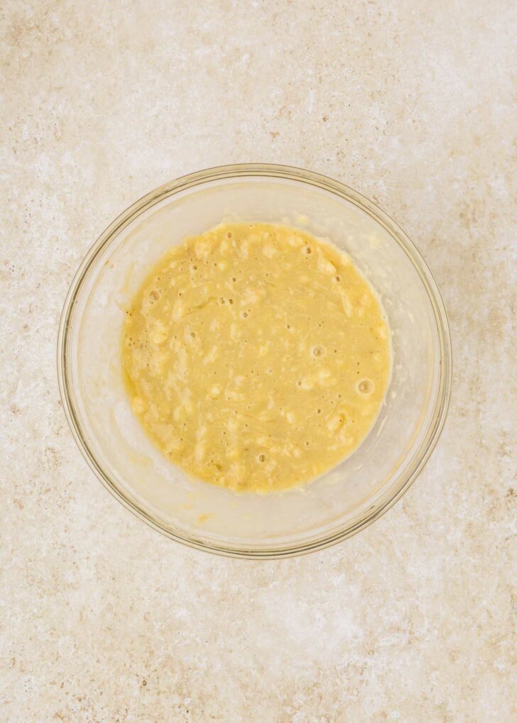 A clear glass bowl filled with a smooth, creamy cake mix banana bread batter rests on a light, speckled countertop. The batter is pale yellow and slightly lumpy, indicating the presence of mashed bananas.