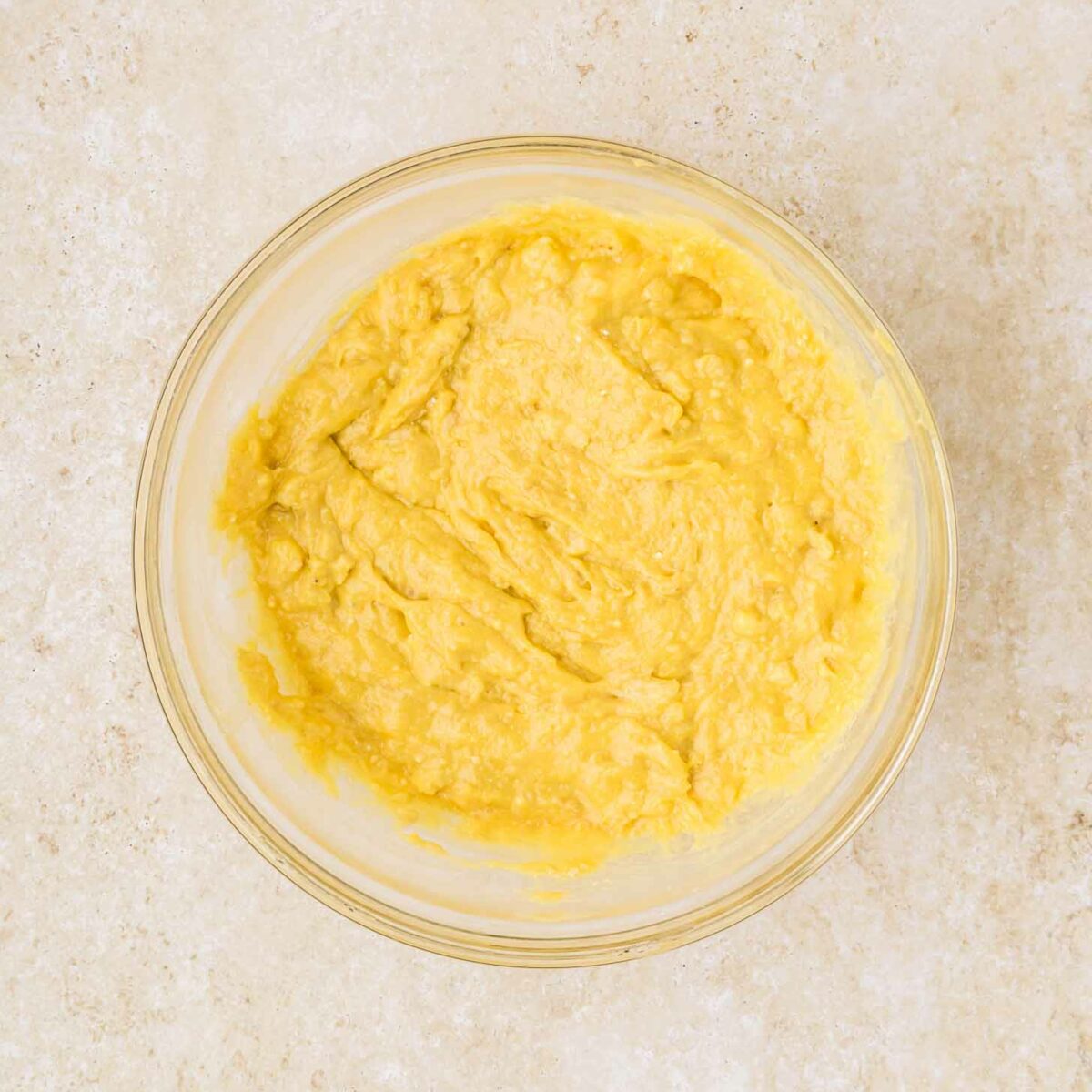A glass bowl filled with creamy yellow batter sits on a textured beige countertop.