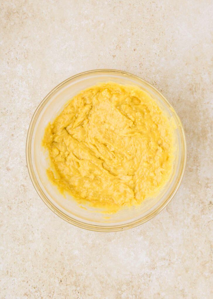 A glass bowl filled with creamy yellow batter sits on a textured beige countertop.
