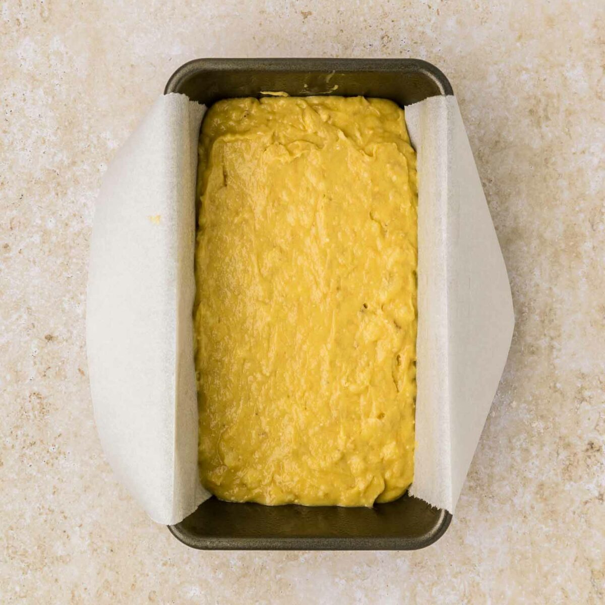 A rectangular metal loaf pan lined with parchment paper is filled with unbaked cake mix banana bread batter. The pan is placed on a beige, textured surface.