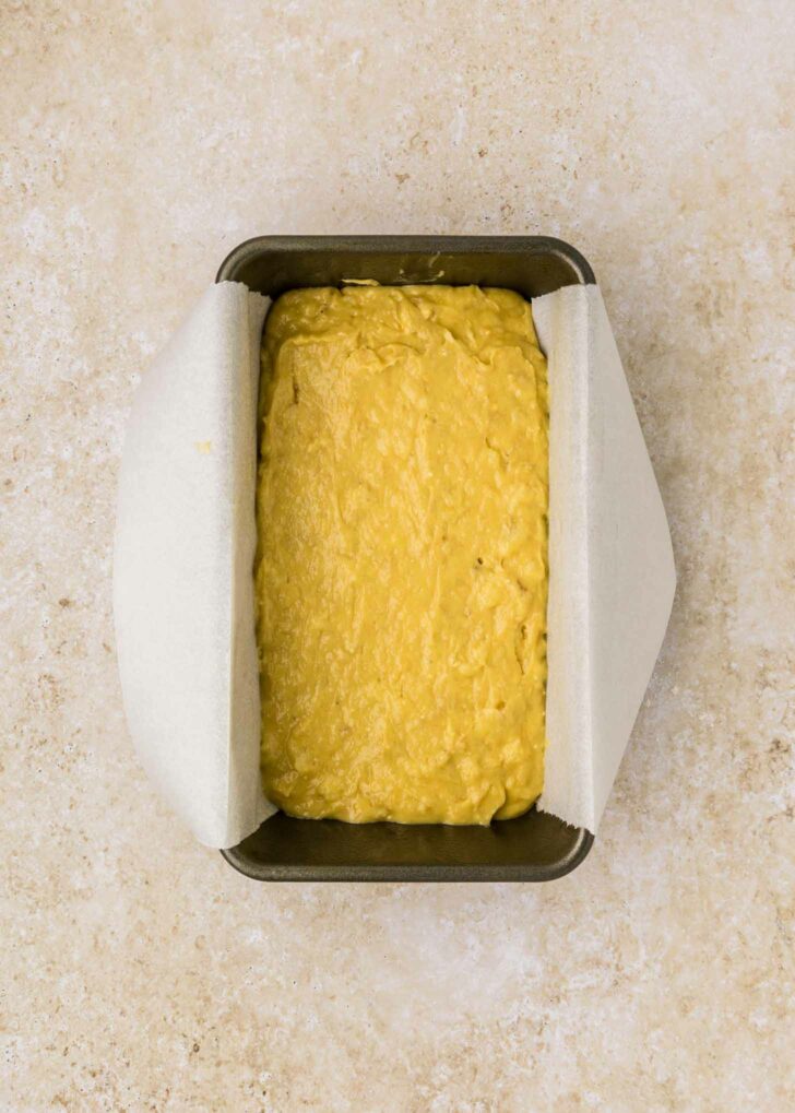 A rectangular metal loaf pan lined with parchment paper is filled with unbaked cake mix banana bread batter. The pan is placed on a beige, textured surface.