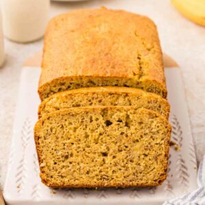 Loaf of sliced banana bread on a white cutting board, accompanied by a glass of milk and a blue-striped cloth. Blurred background includes additional loaf portions and a bunch of bananas.