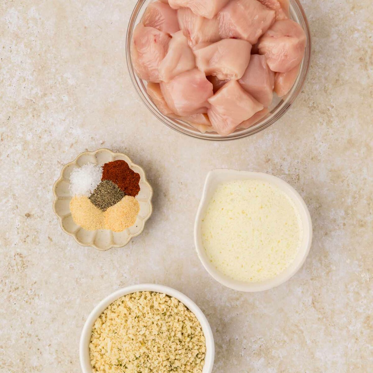 A top-down view of cubed raw chicken in a bowl, surrounded by small bowls containing breadcrumbs, melted butter, and a mix of spices including salt, pepper, and paprika, all on a light-colored surface.