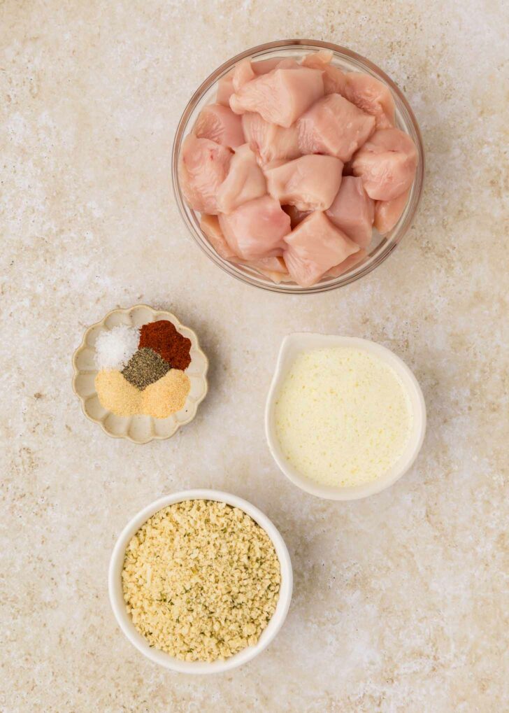 A top-down view of cubed raw chicken in a bowl, surrounded by small bowls containing breadcrumbs, melted butter, and a mix of spices including salt, pepper, and paprika, all on a light-colored surface.