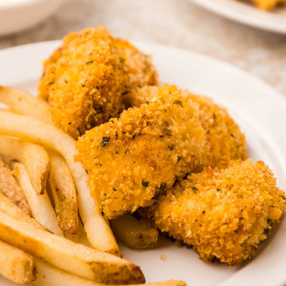 A white plate with golden-brown breaded chicken nuggets and a serving of French fries. A bowl of dipping sauce is blurred in the background.