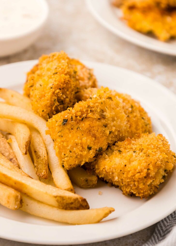 A white plate with golden-brown breaded chicken nuggets and a serving of French fries. A bowl of dipping sauce is blurred in the background.