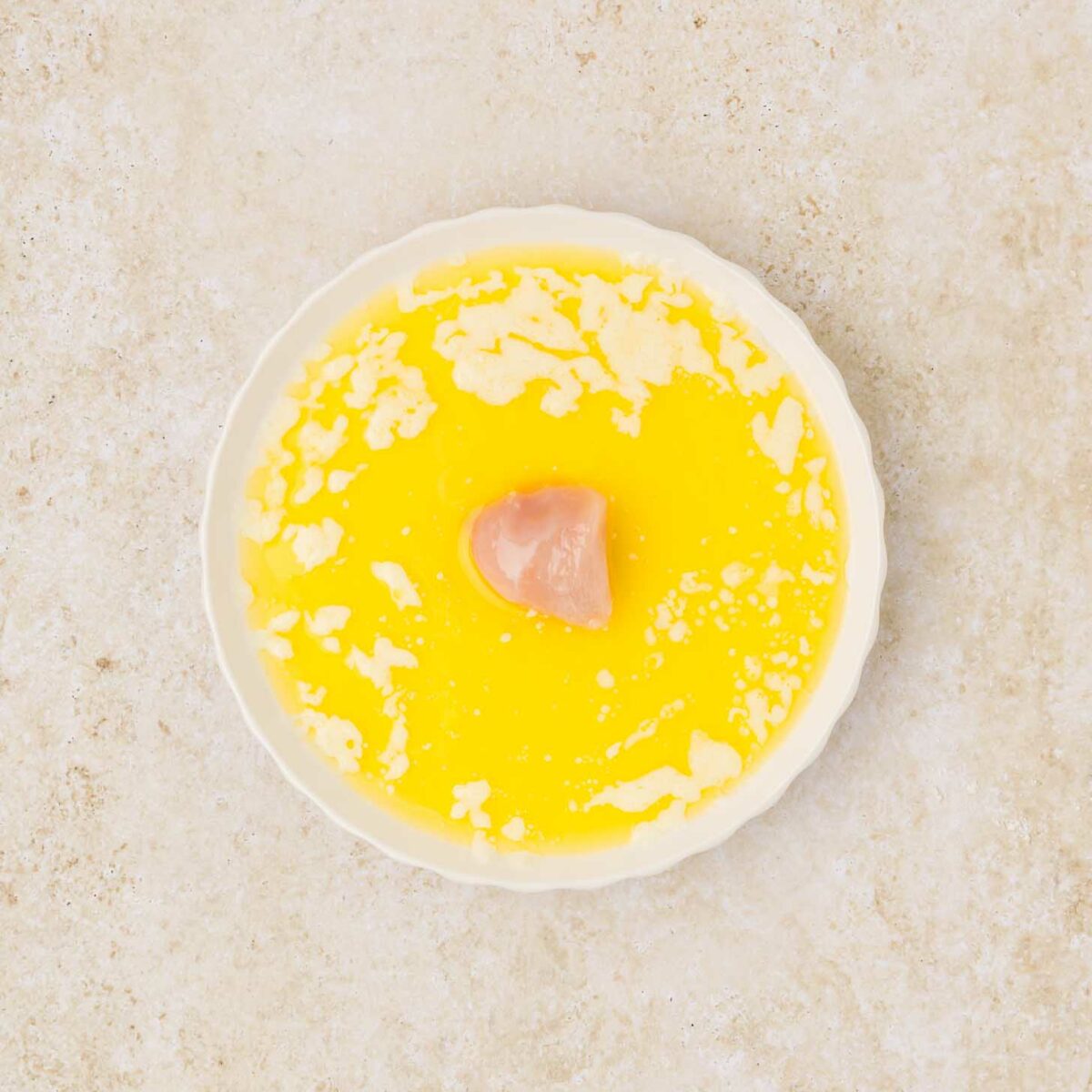 A white round dish contains melted butter, with small solidified bits on the surface and a piece of raw chicken in the center. The background is light and textured.