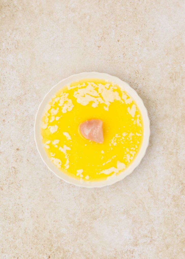 A white round dish contains melted butter, with small solidified bits on the surface and a piece of raw chicken in the center. The background is light and textured.