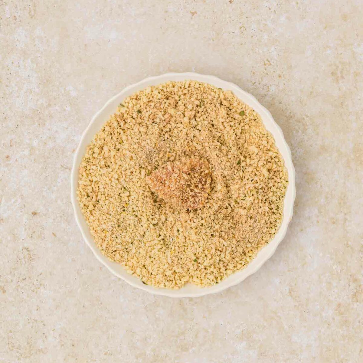 A white plate filled with finely ground breadcrumbs is set against a light beige textured background. The breadcrumbs are light brown and evenly spread on the plate with a piece of chicken in the middle.