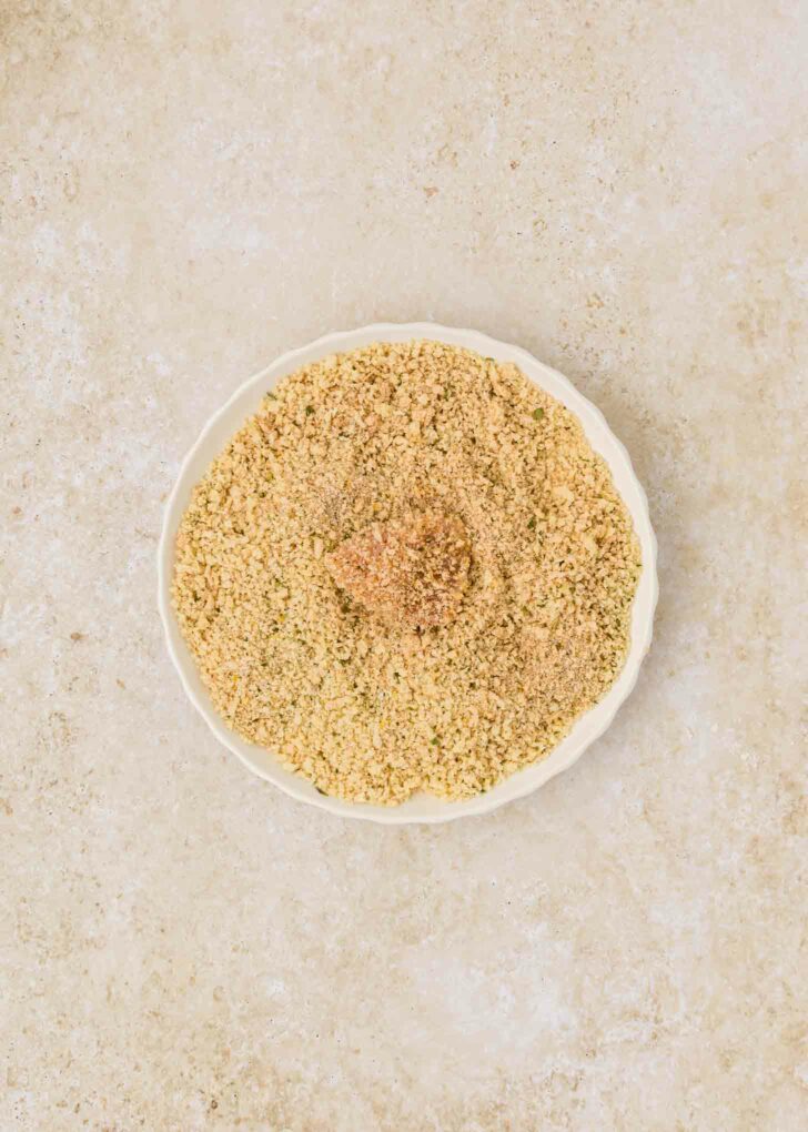 A white plate filled with finely ground breadcrumbs is set against a light beige textured background. The breadcrumbs are light brown and evenly spread on the plate with a piece of chicken in the middle.