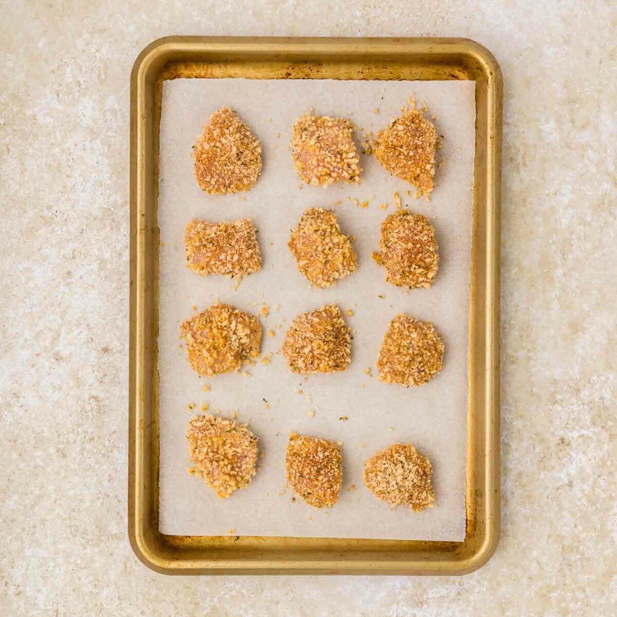 A baking tray lined with parchment paper holds twelve uncooked, breaded chicken nuggets. They are evenly spaced and placed on a light-colored countertop.